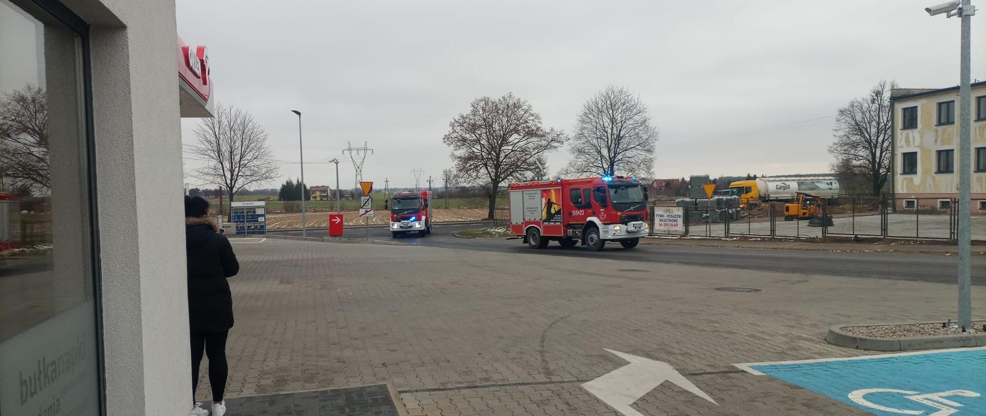 Dwa ciężarowe samochody pożarnicze wjeżdżają na teren stacji Paliw Orlen. Są to ćwiczenia.