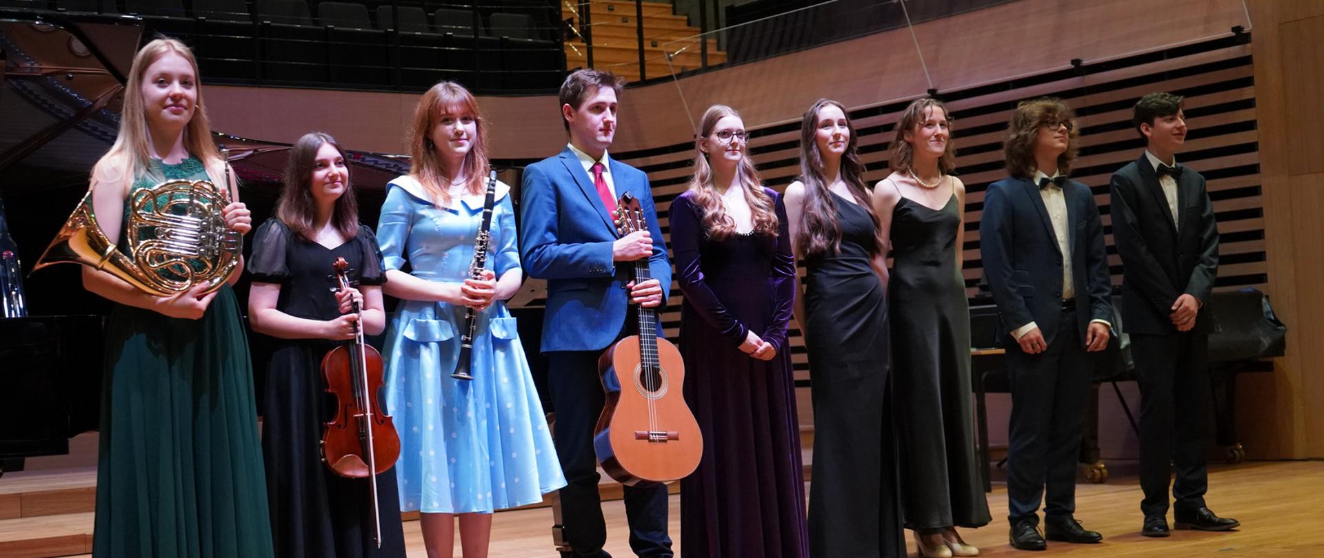 Grupa młodzieży sześciu dziewcząt i trzech chłopców stoi w jednym rzędzie na estradzie sali koncertowej. Osoby po lewej stronie trzymają przed sobą waltornię, altówkę, klarnet i gitarę.