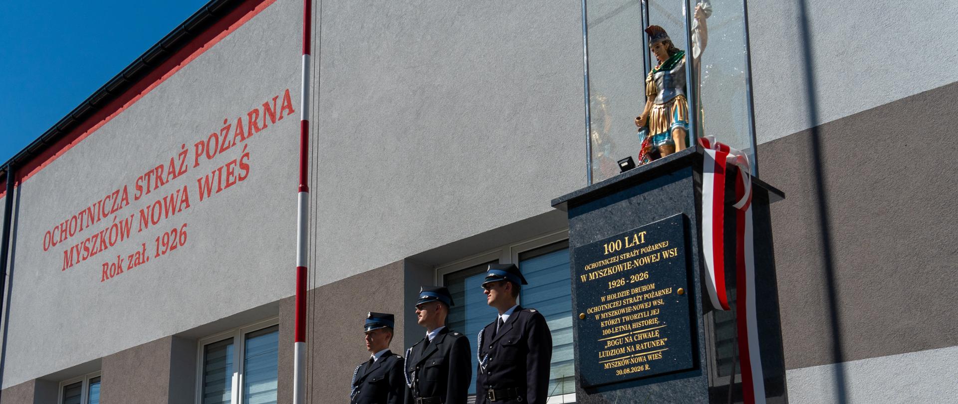 Zdjęcie przedstawia poczet flagowy, w którym stoją druhowie OSP na tle ściany remizy, z prawej stoi postument z tablicą pamiątkową na której stoi figurka Św. Floriana