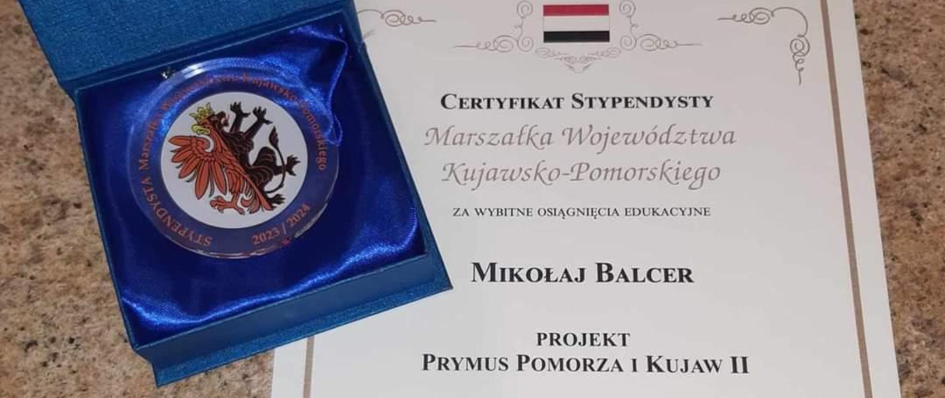 list gratulacyjny dla ucznia biała kartka z czarnymi napisami po lewej stronie medal z narysowanym czerwono czarnym orłem w niebieskim opakowaniu