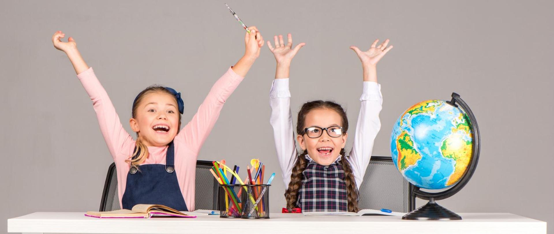 Na zdjęciu znajdują się dwie dziewczyny siedzące przy biurku z uniesionymi do góry rękoma. Na biurku jest globus, kredki i zeszyty.
