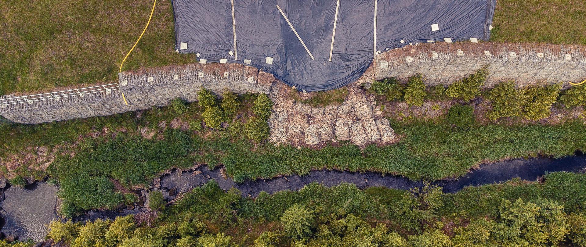 To zdjęcie przedstawia widok z lotu ptaka na plac budowy i miejsce naprawy rozmytej drogi. Widać odcinek drogi, który został zabezpieczony barierkami i plandekami, co wskazuje na trwające prace. Obok drogi znajduje się osunięta kamienna ściana, częściowo zrujnowana, która jest jednym z głównych celów prac naprawczych. Poniżej ściany płynie rzeka. 