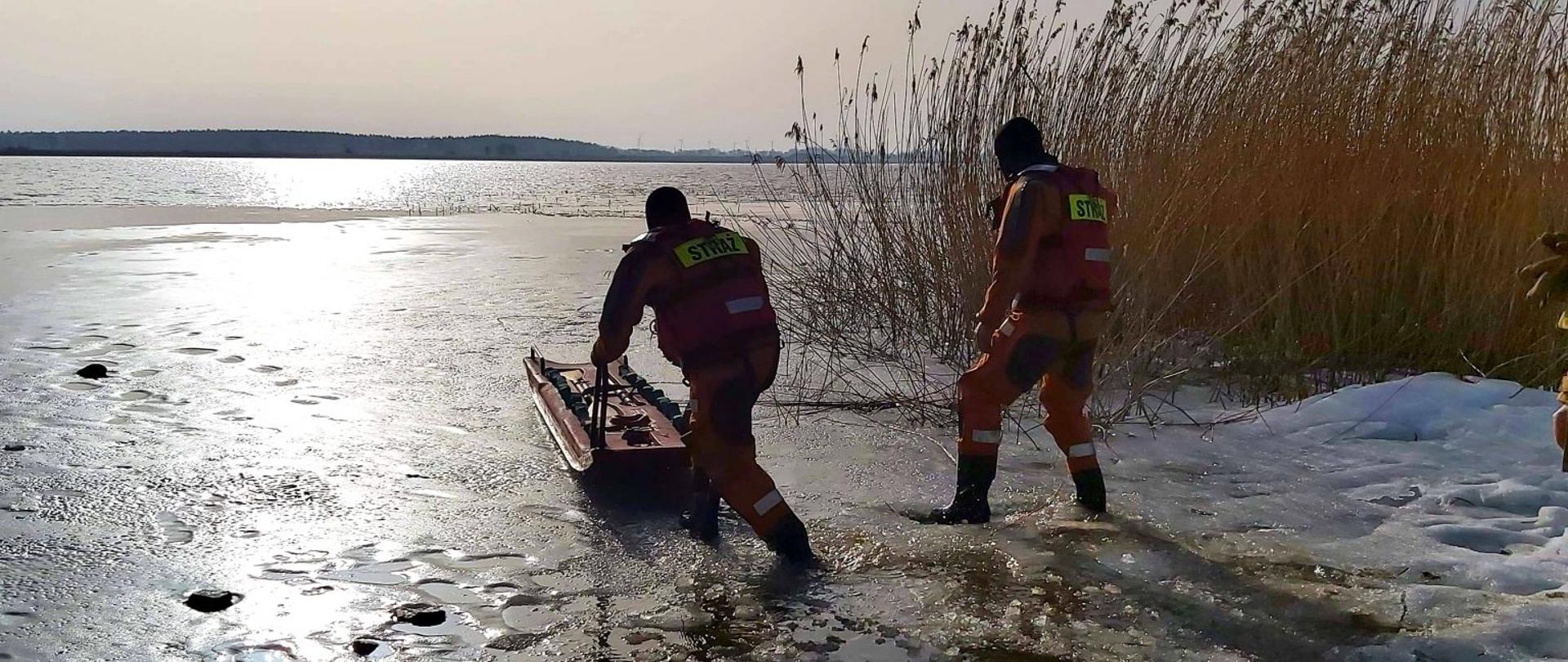 zdjęcie przedstawia dwóch strażaków w skafandrach z saniami lodowymi wchodzących na częściowo zamarznięty akwen.