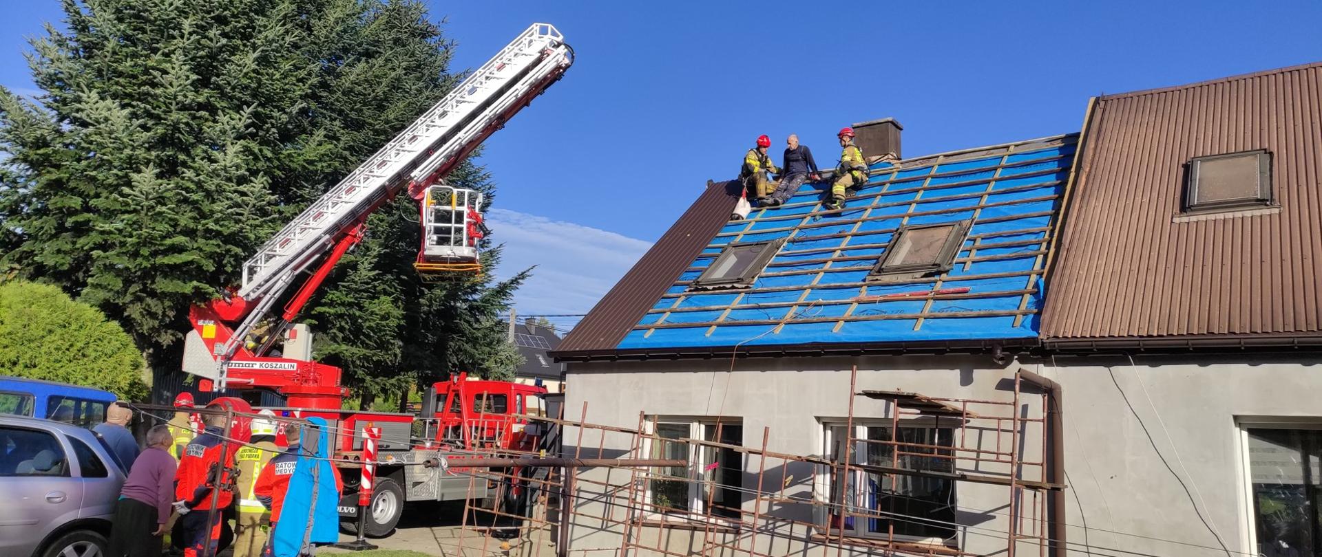Na zdjęciu widoczny budynek mieszkalny 2 kondygnacyjny. Na dachu budynku znajduje się osoba poszkodowana i dwóch strażaków ratowników. Obok budynku znajduje się podnośnik hydrauliczny którym ewakuowano osobę poszkodowaną oraz pozostali ratownicy.