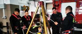 Garaż Jednostki Ratowniczo-Gaśniczej, strażacy uczestniczący w zajęciach doskonalenia zawodowego z ratownictwa wysokościowego. Wykorzystują trójnóg oraz sprzęt wysokościowy i z\ochrony osobistej 