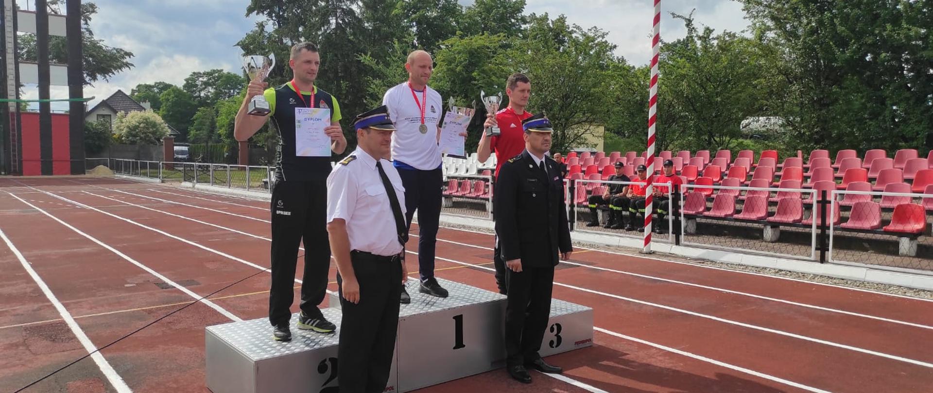 Zdjęcie wykonano na stadionie lekkoatletycznym. Na podium, ustawionym na bieżni, stoi trzech zawodników w strojach sportowych trzymających dyplomy i puchary. Przed podium stoi dwóch strażaków w umundurowaniu wyjściowym.