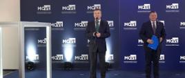 Minister Wieczorek stoi i mówi do mikrofonu na stojaku, obok niego stoi trzymający niebieską teczkę mężczyzna w garniturze, za nim granatowa ścianka z napisem MOVE.