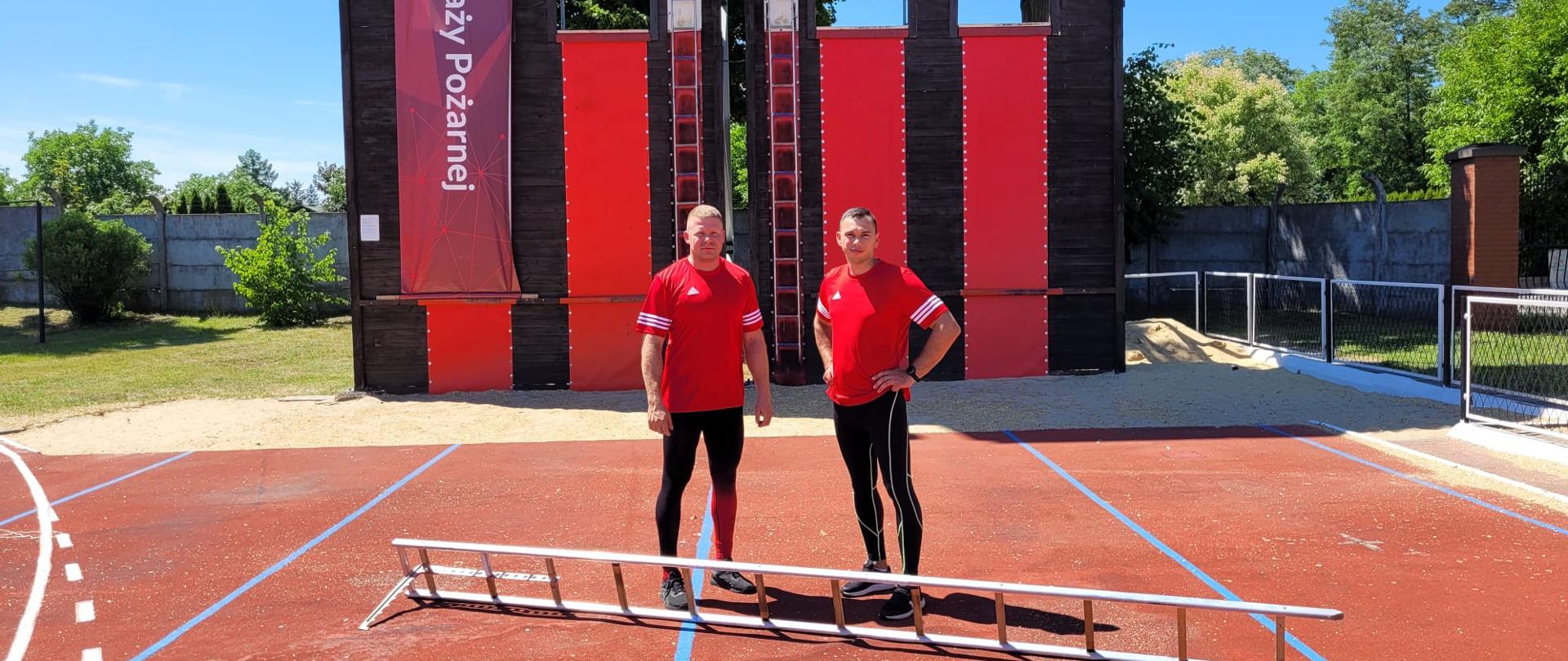 Zdjęcie przedstawia strażaków w koszulkach koloru czerwonego. Strażacy stoją na tle ściany wspinalni, na podłożu leży drabina hakowa.