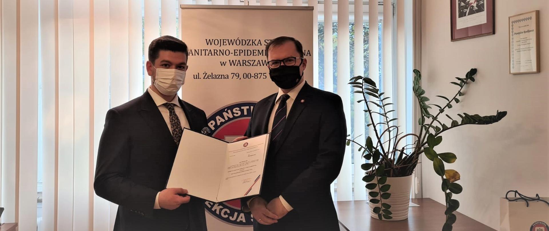 dwaj mężczyźni w maseczkach ochronnych, mężczyzna z lewej trzyma w ręku powołanie na inspektora sanitarnego w Płocku