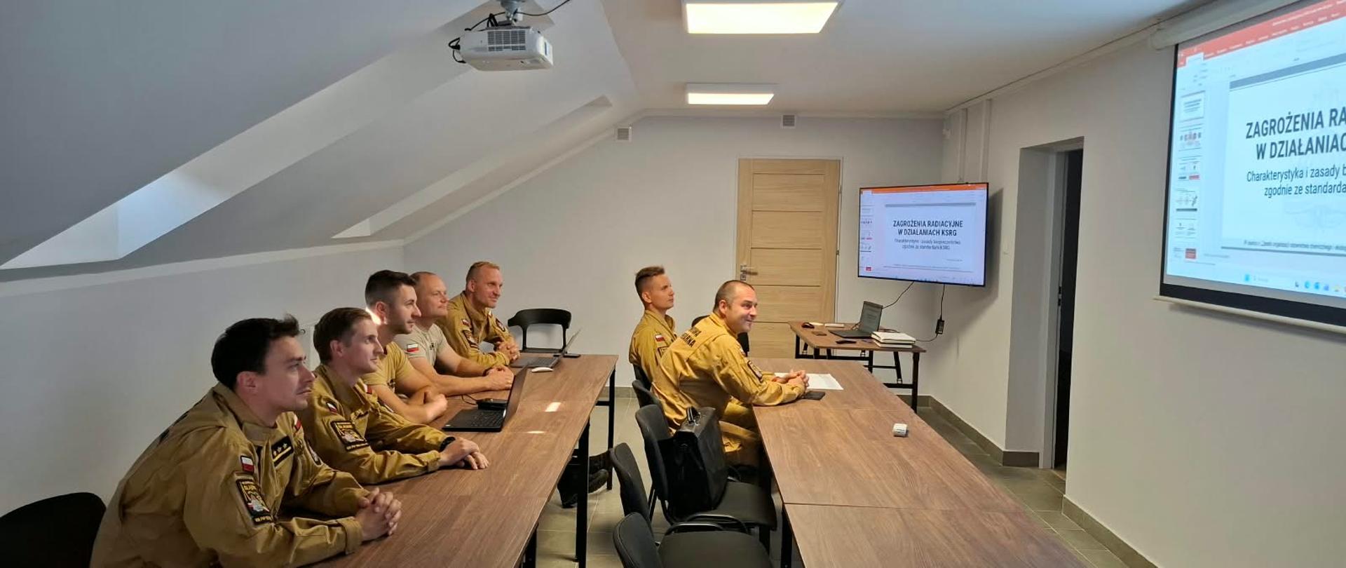 Na zdjęciu widać grupę strażaków Państwowej Straży Pożarnej podczas szkolenia w sali wykładowej. Funkcjonariusze w umundurowaniu służbowym w kolorze piaskowym, siedzą przy stołach i słuchają prowadzącego. Na ekranach wyświetlana jest prezentacja dotycząca zagrożeń radiacyjnych i zasad działania w sytuacji zagrożenia. Sala wyposażona jest w projektor, monitory oraz sprzęt komputerowy.