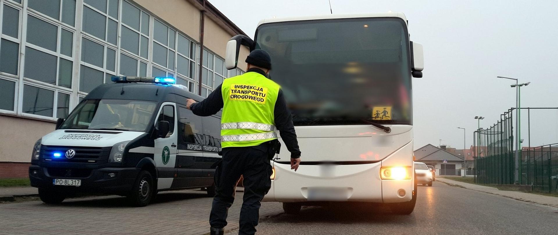 Na pierwszym planie: umundurowany inspektor wielkopolskiej Inspekcji Transportu Drogowego. W tle (od lewej): oznakowany furgon wielkopolskiej Inspekcji Transportu Drogowego i stojący obok kontrolowany autobus.