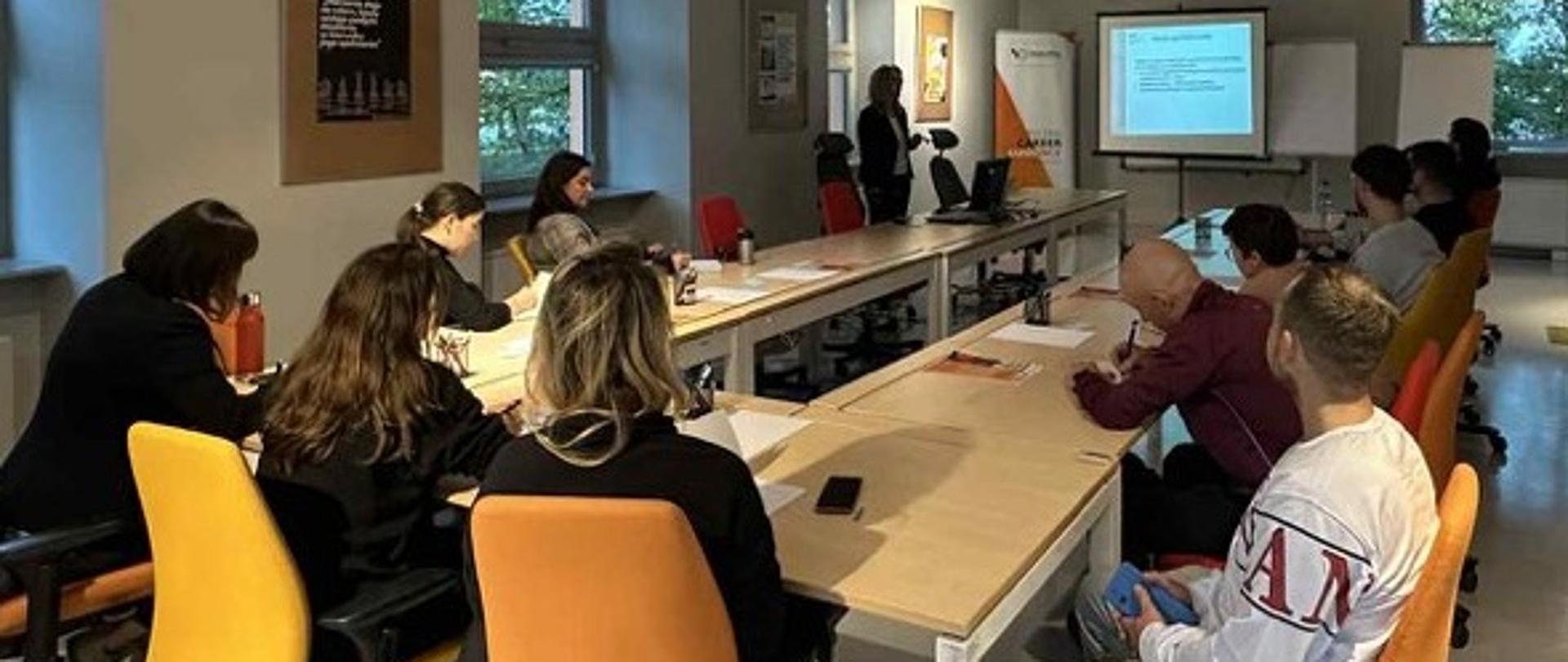 Grupa osób siedzi w sali przy stołach i słucha wykładu o podatkach