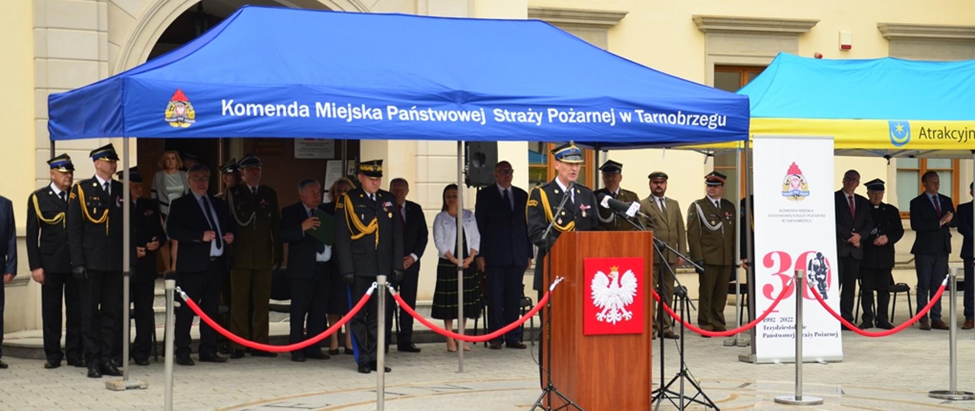 Na dziedzińcu Zamku Dzikowskiego przed mównicą stoi strażak, który przemawia do zgromadzonych, za nim pod pawilonem widoczni zaproszeni gości, wśród których stoją oficerowie Państwowej Straży Pożarnej, Wojska Polskiego oraz innych służb mundurowych. 