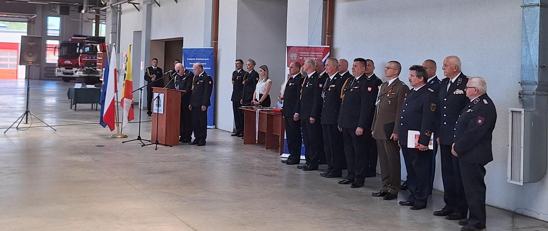 umundurowani strażacy stojący w szeregu w pomieszczeniu hali garażowej, po lewej stronie mównica, nad strażakami baner z napisem : dzień strażaka 