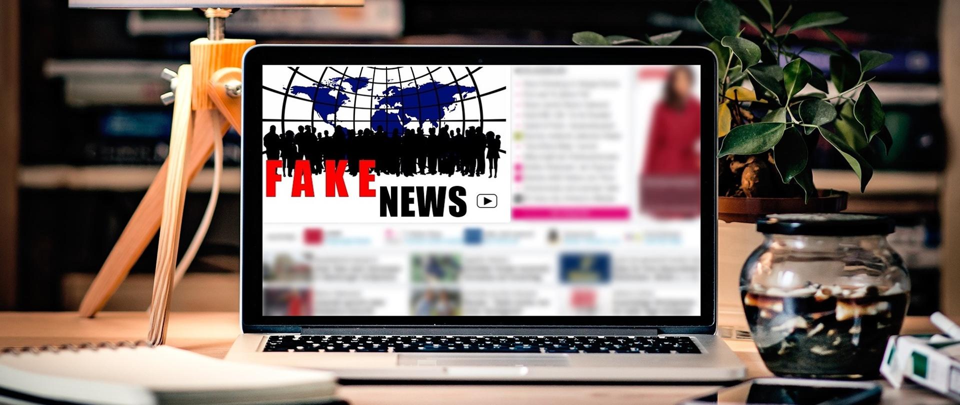 Zdjęcie przedstawia biurko, na którym stoi laptop. Na jego ekranie widnieje napis Fake News. W tle zdjęcia znajdują się regał z książkami. Ponadto, po lewej stronie na biurku widać lampę z drewnianymi nogami oraz jasnym, okrągłym abażurem. Po prawej stronie paczka papierosów oraz słoik. Za słoikiem stoi roślinka domowa. 
