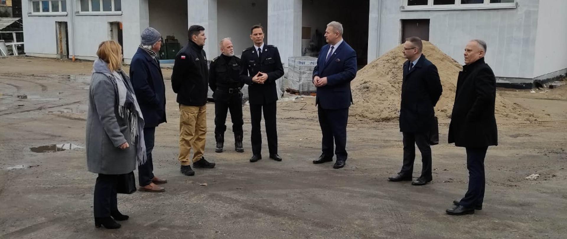 Delegacja wizytująca plac budowy ustawiona na placu zewnętrznym komendy omawia postęp prac. W tle budynek garażowy.