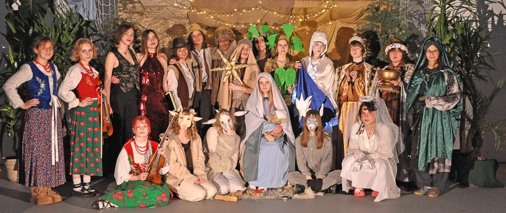 Na zdjęciu jest grupa ludzi przebranych za postacie z jasełek bożonarodzeniowych. Zdjęcie przedstawia scenę narodzin Jezusa. Osoby na zdjęciu są ubrane w stroje reprezentujące Maryję, Józefa, Trzech Króli, pasterzy i aniołów. Trzej Królowie są ubrani kolorowo i mają na głowach korony; jeden z nich trzyma gwiazdę. Inne postacie są ubrane w tradycyjne stroje, reprezentujące pasterzy lub mieszkańców Betlejem. W tle znajduje się dekoracja przypominająca stajnię. 