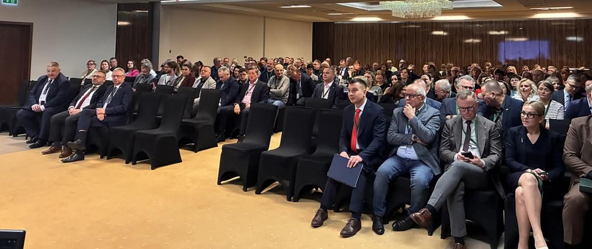 Na czarnych krzesłach siedzi duża grupa ludzi, w dużej sali