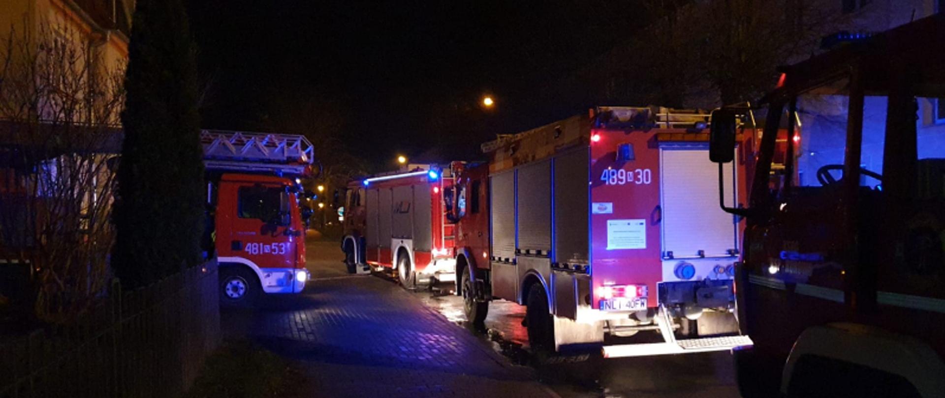 Zdjęcie przedstawia 3 samochody pożarnicze zaparkowane równolegle do drogi . W wewnętrznej drodze zaparkowany jest podnośnik hydrauliczny. 