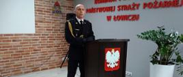 na zdjęciu komendant powiatowy PSP w Łowiczu mł. bryg. Arkadiusz Makowski, który stojąc przy mównicy wygłasza przemówienie podczas ceremonii wręczenia odznaczeń i awansów w stopniach dla funkcjonariuszy tutejszej komendy. Komendant ubrany jest w mundur galowy ze sznurem stojący przy mównicy na której widnieje godło państwowe, obok zielony kwiat w białej donicy, w tle ściana z imitacją brązowej cegły na niej logo PSP. zdjęcie w formacie panorama.
