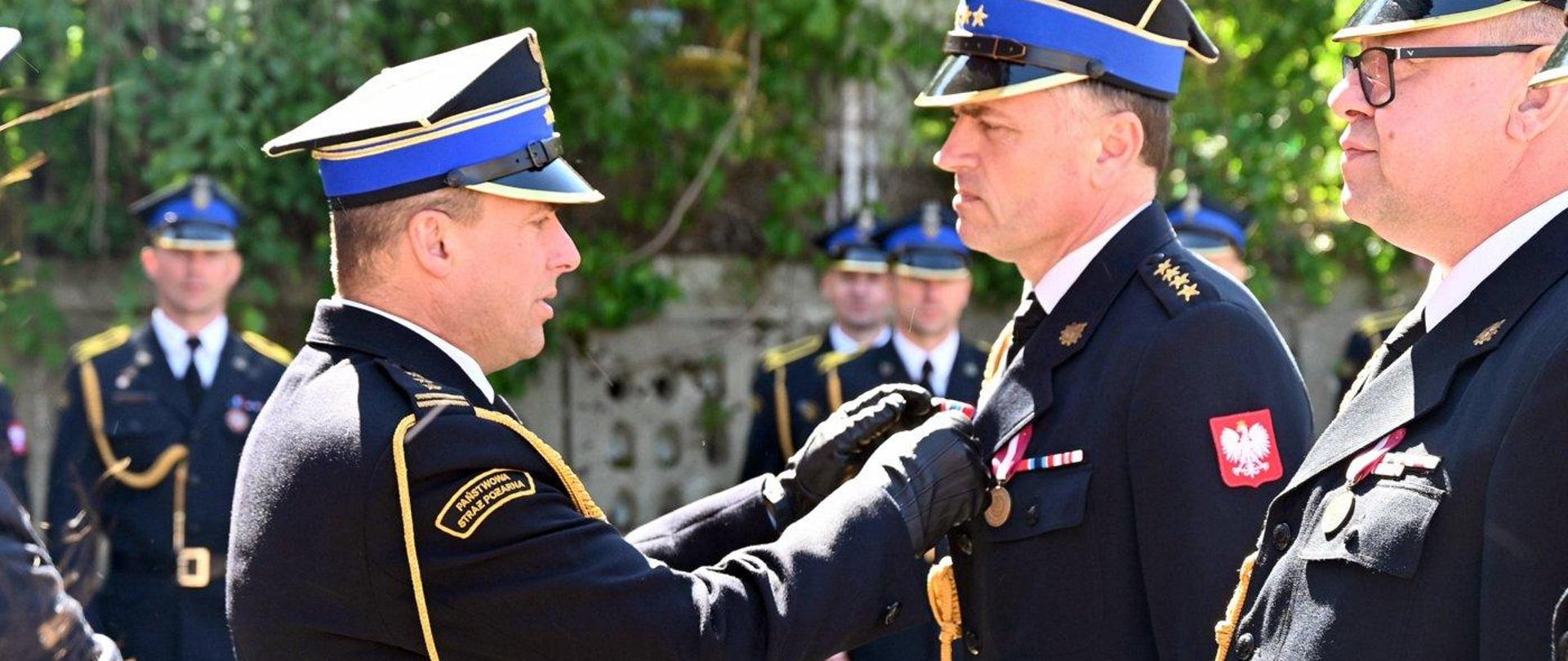 Kolorowa fotografia wykonana pogodny dzień na zewnątrz. Przedstawia Podkarpackiego Komendanta Wojewódzkiego PSP podczas wręczania odznaczeń na uroczystości Dnia Strażaka.