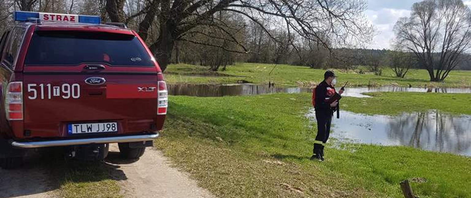 Rzeka Czarna Włoszczowska, strażak ratownik ubrany w ubranie koszarowe, zabezpieczony kamizelką ratunkową, a brzegu rzeki przeprowadza kontrolę poziomu wody, Na drodze zaparkowany jest samochód pożarniczy.