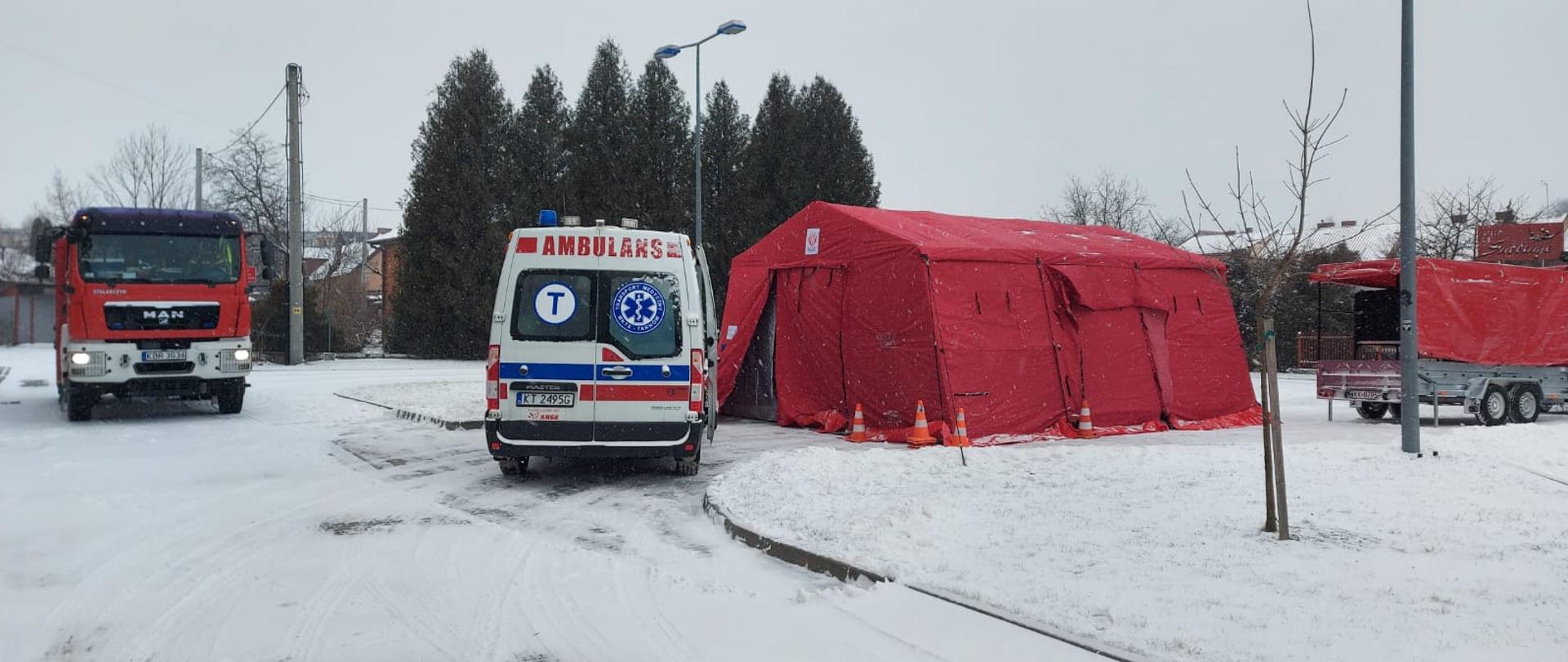Czerwony namiot do poboru próbek obok ambulans i pojazd straży pożarnej. Teren zaśnieżony.
