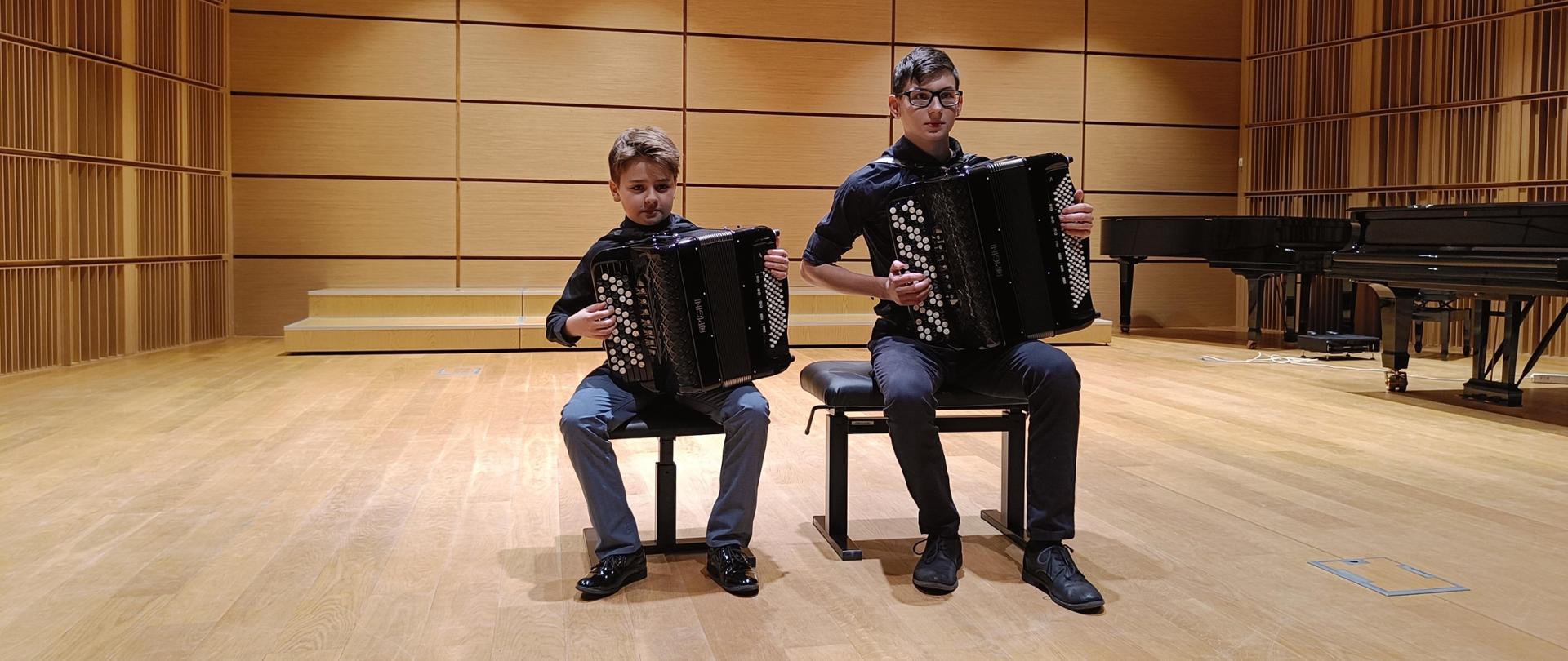 Dwóch uczniów trzymających akordeony na scenie sali koncertowej. Z tyłu, po prawej stronie widać fragmenty dwóch fortepianów.