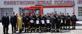 Zakończenie szkolenia podstawowego Ochotniczych Straży Pożarnych. Na tle jednostki straży pożarnej ustawiony pojazd ratowniczy oraz pododdział strażaków w umundurowaniu koszarowym. Strażacy trzymają w dłoniach zaświadczenia kończące szkolenie. 