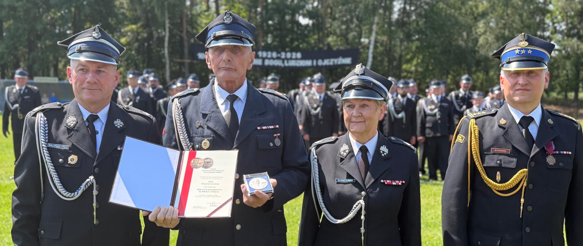 Mundurowi pozują w rzędzie na tle zielenicy i zgromadzonych w tle strażaków. Jeden ze starszych strażaków prezentuje otwarty dyplom pamiątkowy oraz pamiątkowy medal w etui.