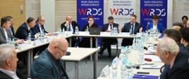 Widok na salę z żółtymi ścianami, przy ustawionych w czworokąt stołach siedzą ludzie, u szczytu stołu siedzi minister Czarnek, kobieta w niebieskiej marynarce, kobieta w czarnej marynarce i dwóch mężczyzn w garniturach, za nimi banery z napisem WRDS i zasłonięte żaluzjami okna.