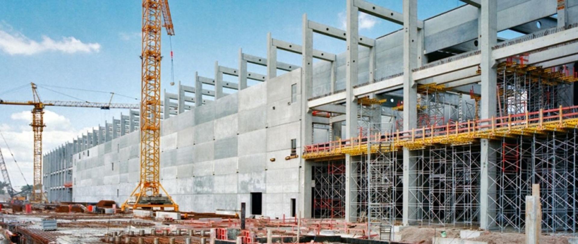 Budowa wielopiętrowego obiektu z dźwigami, rusztowaniami i rozstawionym sprzętem na placu budowy.