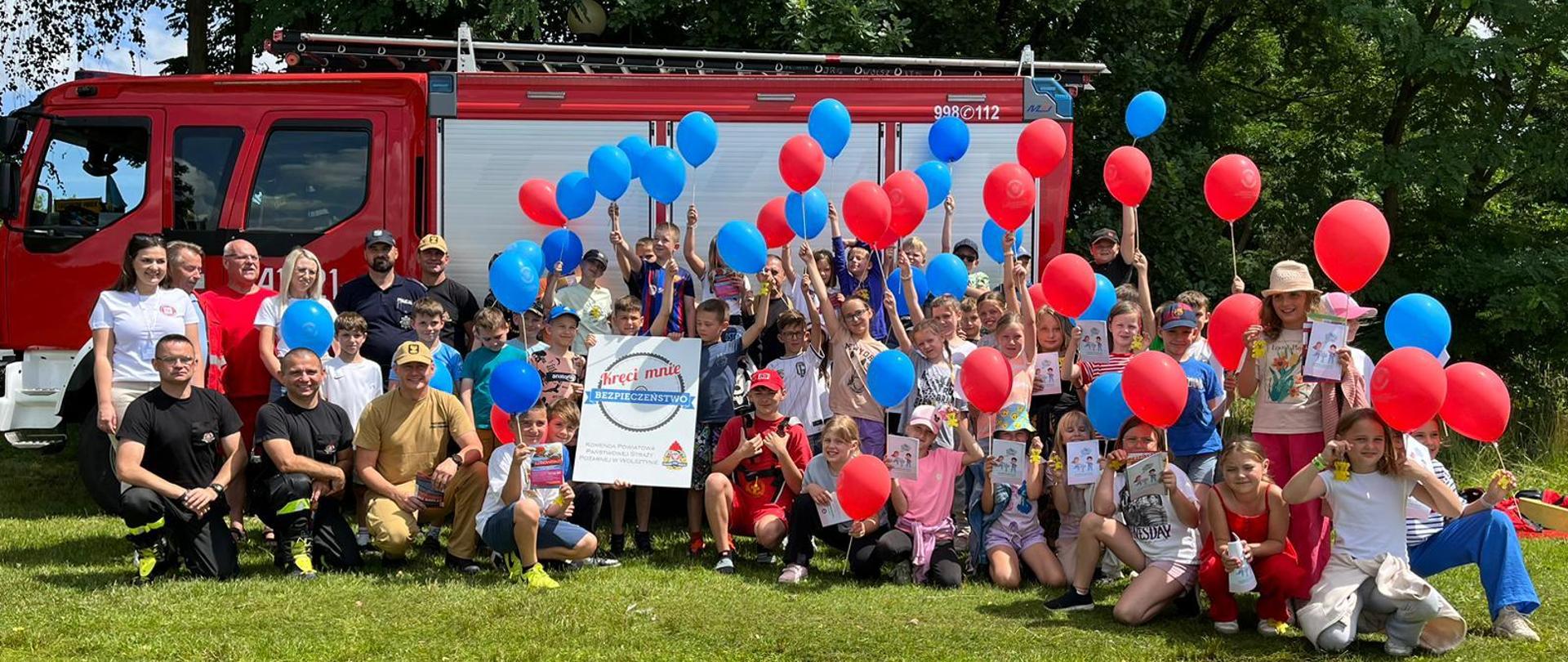 Na zdjęciu uczestnicy akcji z niebieskimi i czerwonymi balonami w ręku wraz ze strażakami ustawieni przed samochodem strażackim