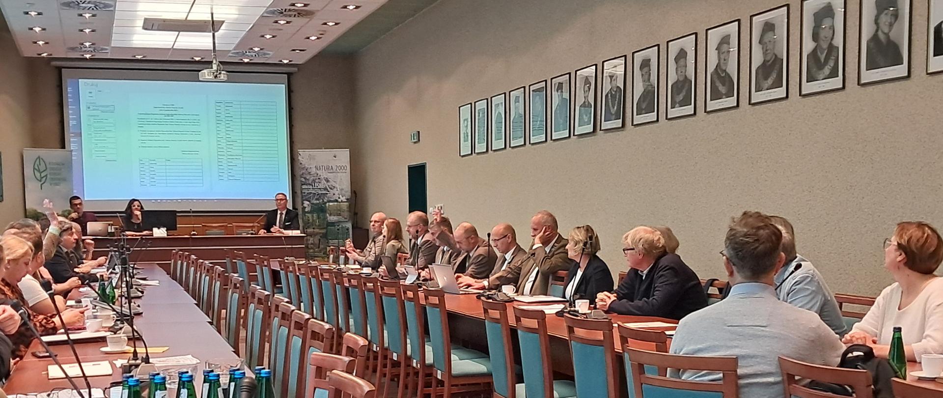 sala, ludzie siedzący za sołami, na ścianie obraz z rzutnika, wyświetlone tabelki