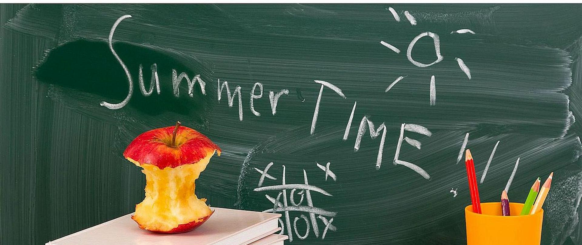 Plakat przedstawia informacje dotyczące zakończenia roku szkolnego. W centralnej części plakatu znajduje się szkolne biurko z kredkami, książkami, kubkiem i ogryzkiem jabłka leżącymi na nim. W tle tablica szkolna z napisem "Summer time".