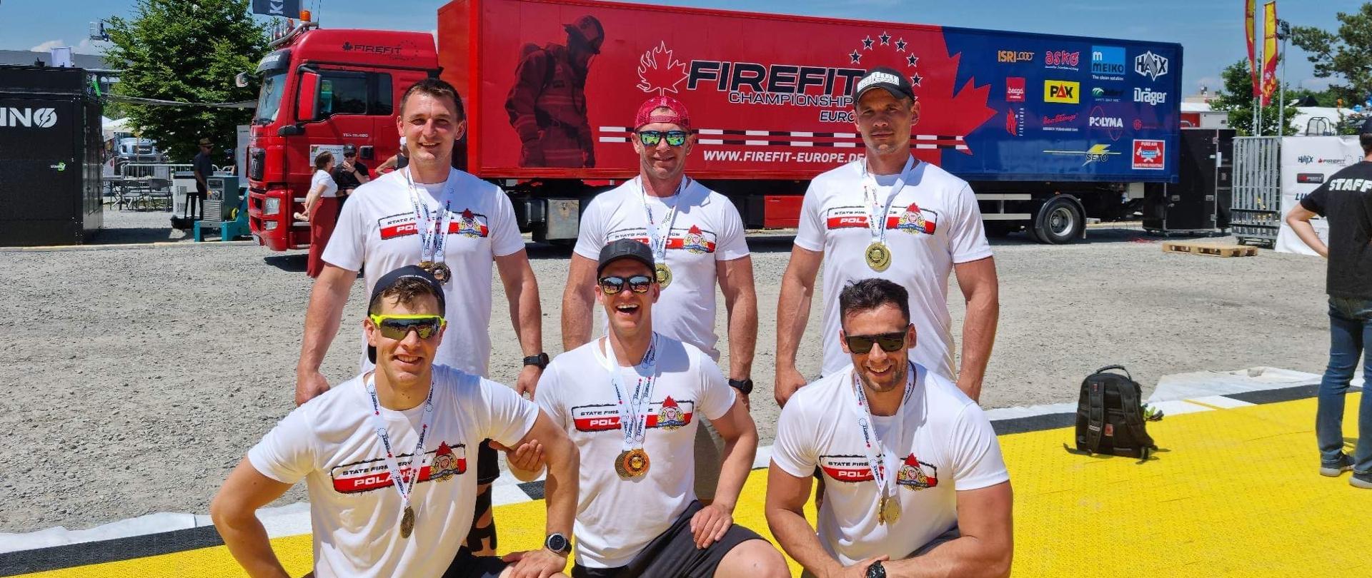 5 mężczyzn w koszulkach sportowych z medalami zawieszonymi na szyjach, za nimi ciężarówka w kolorach czerwieni i granatu z napisem FIREFIT