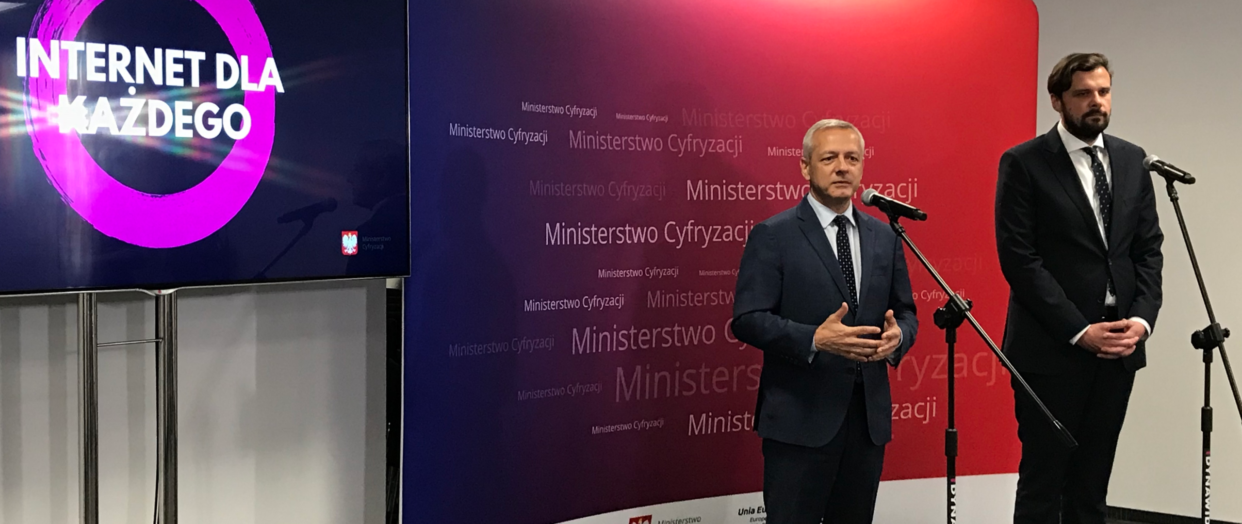 Minister Marek Zagórski i dyrektor Wojciech Szajnar stoją na tle ściany z napisami: Ministerstwo Cyfryzacji. Obok ścianki stoi telewizor. Przed ministrem i dyrektorem ustawione są mikrofony.