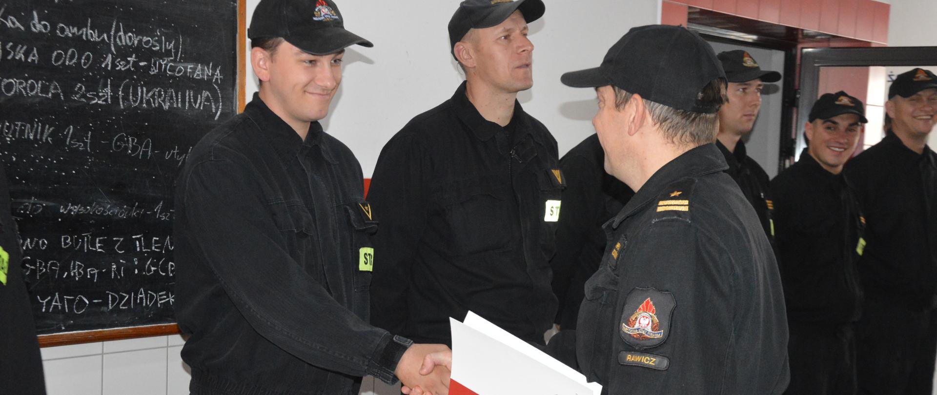 Wręczenie decyzji nagrodowej. Garaż Komendy Powiatowej PSP w Rawiczu. Na przeciwko siebie stoją: komendant powiatowy oraz nagrodzony strażak. Komendant wręcza decyzję o przyznaniu nagrody , ściskając jednocześnie dłoń wyróżnionego strażaka. W tle tablica, z prawej strony strażacy ze zmiany służbowej JRG.