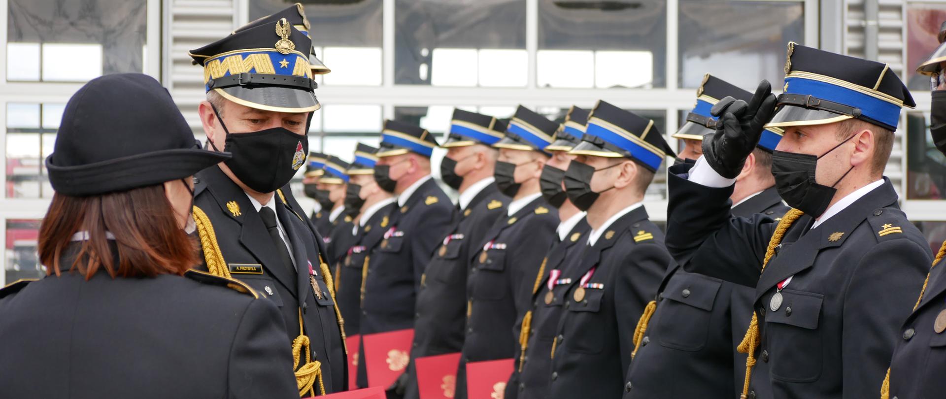 Zastępca Komendanta Głównego PSP wręcza akty nadania stopni służbowych, po prawo ustawieni w szeregu funkcjonariusze PSP w mundurach galowych, w lewej dłoni trzymają czerwone teczki 