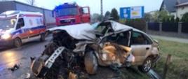 Zdjęcie przedstawia zniszczony w czasie wypadku samochód osobowy. W tle widoczne pojazdy pogotowia ratunkowego oraz Państwowej Straży Pożarnej