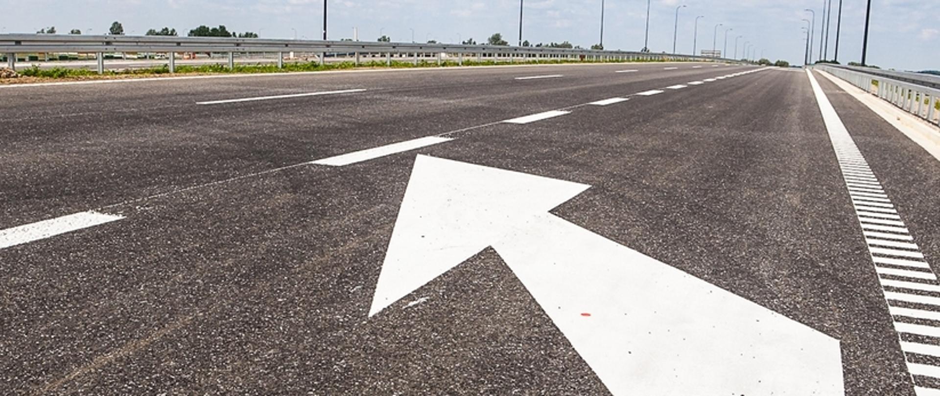 Zdjęcie przedstawia fragment autostrady A4. W centralnej części kadru widoczna jest szeroka jezdnia z oznakowaniem poziomym w postaci białej strzałki kierunkowej wskazującej jazdę na wprost. Po obu stronach drogi rozmieszczone są metalowe bariery ochronne, które zapewniają bezpieczeństwo użytkowników drogi. W tle można dostrzec dalszy odcinek autostrady, latarnie oraz otaczający krajobraz, prawdopodobnie składający się z pól lub łąk.