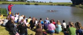 Dzieci ze szkoły podstawowej siedzą na trawie w pobliżu stawu. Obok nich stoi strażak ubrany w skafander do pracy w wodzie. Funkcjonariusz demonstruje w jaki sposób poprawnie należy używać rzutki ratowniczej. Drugi strażak znajduje się w wodzie, udaje osobę która się topi. W tle pola i drzewa wokół stawu.