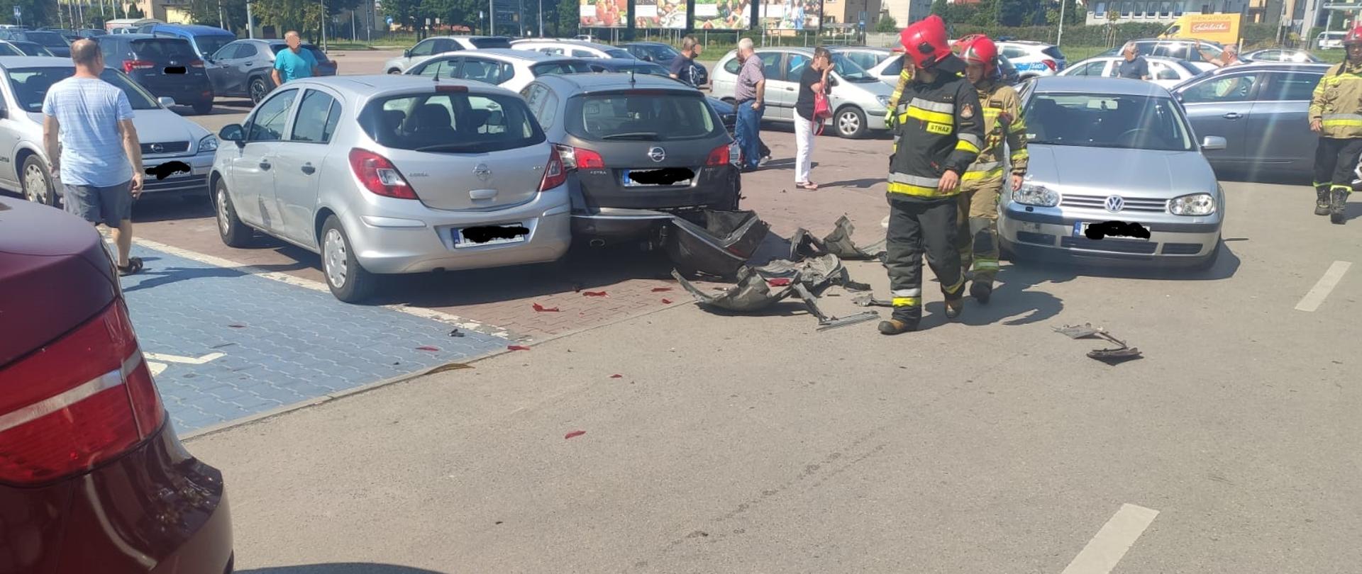 Na zdjęciu widzimy uszkodzone samochody osobowe po kolizji na parkingu sklepu. Widać rozsypane elementy karoserii oraz strażaków przechodzących obok pojazdów. W tle widać zaparkowane samochody na parkingu oraz przechodniów.