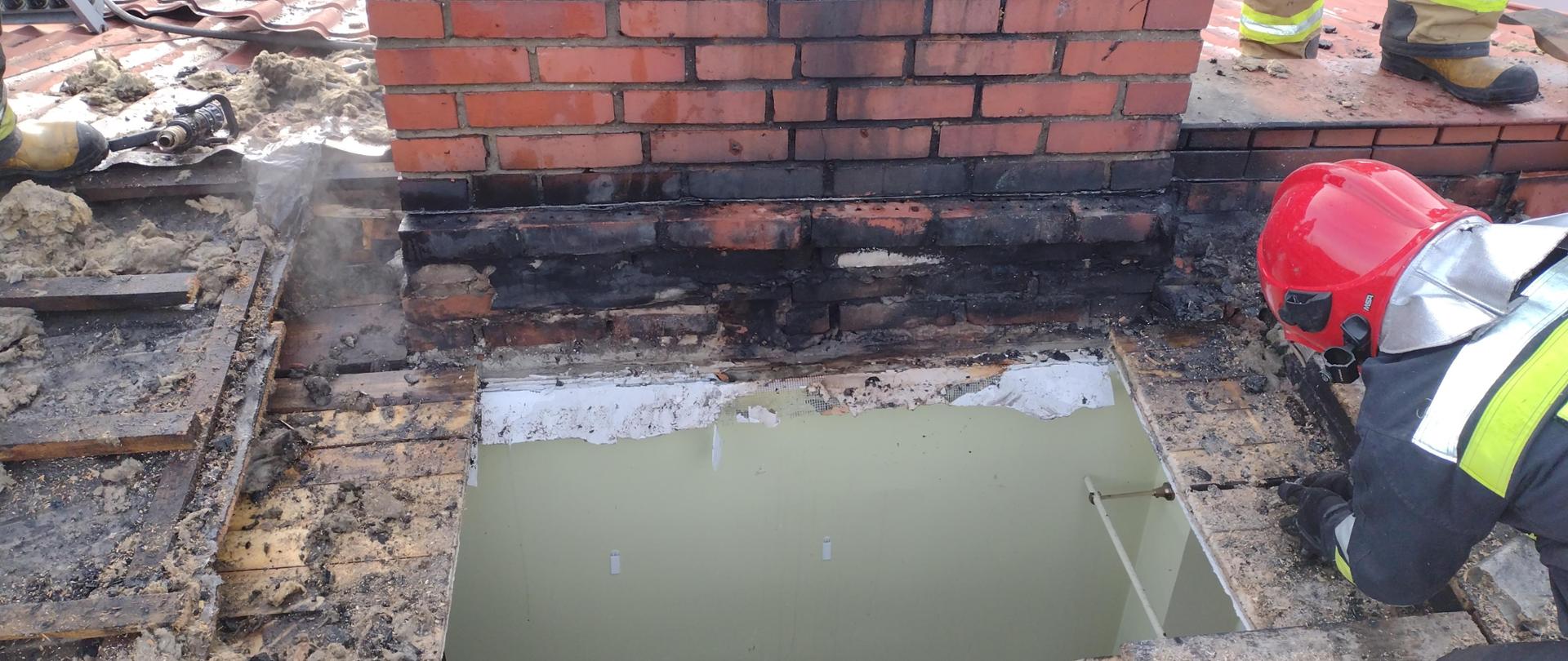 Na zdjęciu widać dwóch strażaków, którzy znajdują się na dachu budynku w którym doszło do pożaru dachu. Strażacy wykonują otwór w dachu by dogasić tlące się elementy konstrukcji dachu.