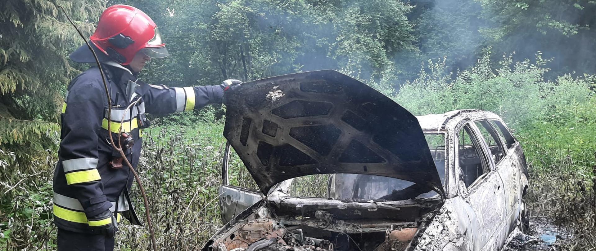 Strażak obok spalonego samochodu w lesie trzyma otwartą klapę silnika.