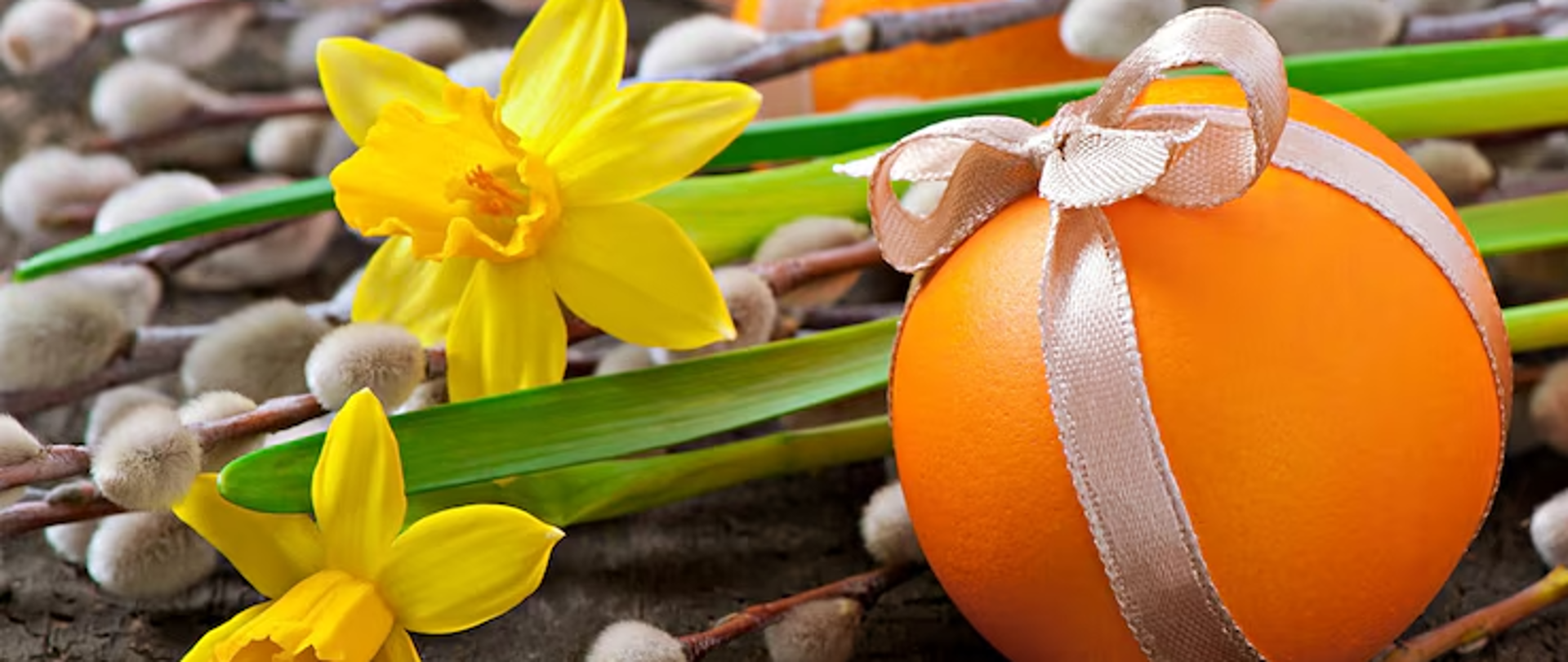 Ułożone poziomo białe bazie. Pomiędzy baziami po prawej stronie leżą 2 żółte pisanki przewiązane beżową wstążką zawiązaną u góry na kokardkę. Między pisanki włożone są 2 żółte kwiaty żonkili, których a zielone łodygi oddzielają pisanki. 