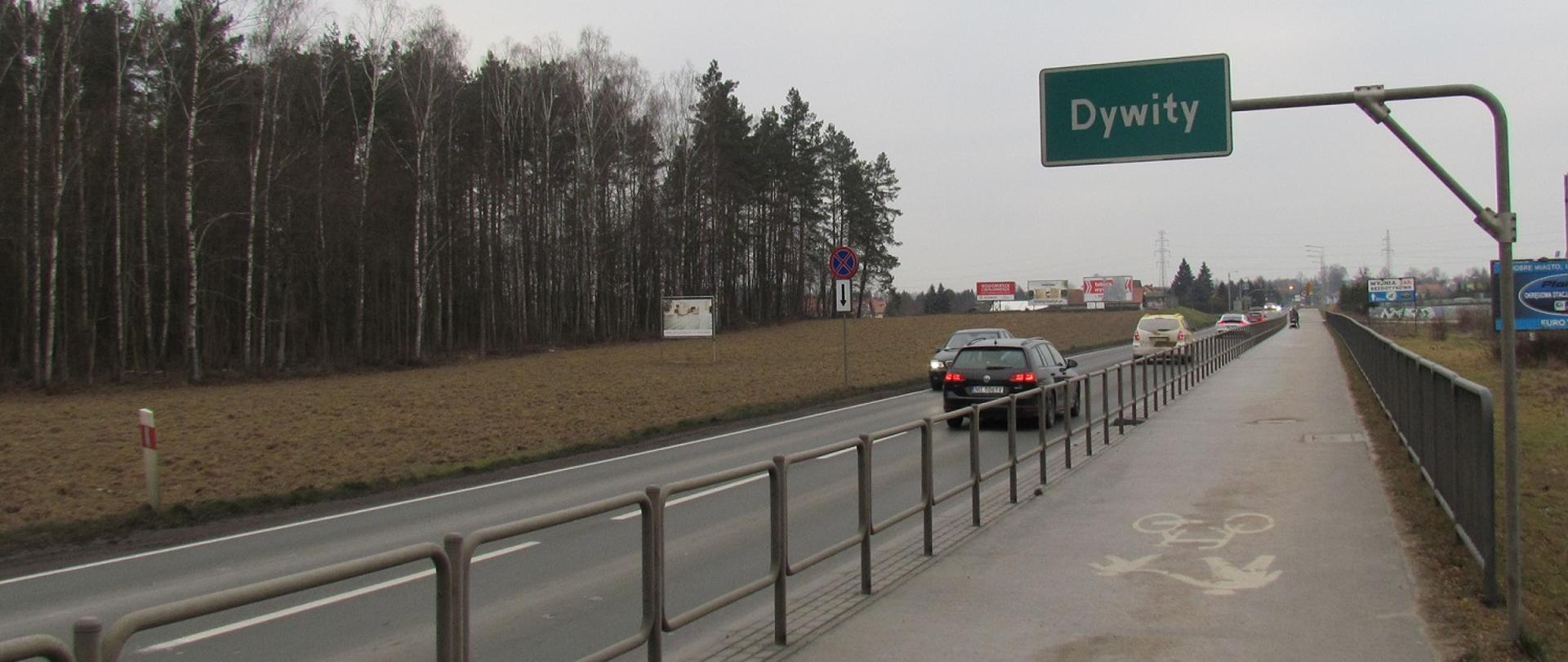 Asfaltowa droga, obok za barierkami ścieżka rowerowa. Na jezdni sznur samochodów. Nad drogą tablica z nazwą miejscowości. 