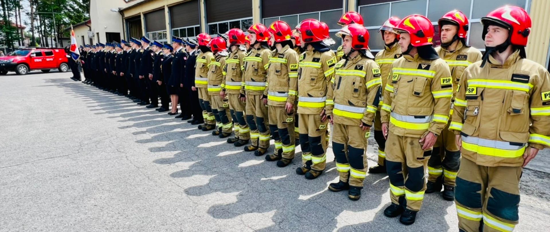 Na tle budynku straży stoi szereg strażaków umundurowanych w strażackie umundurowaniu specjalne oraz umundurowanych w stroje wyjściowe.