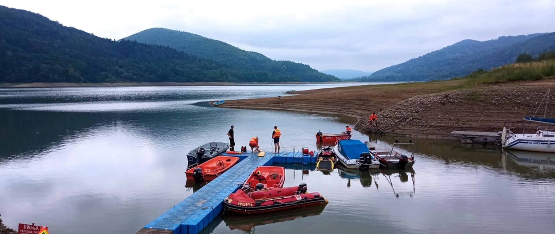 Na zdjęciu widoczny zalew Klimkówka oraz pomost na których stoją strażacy uczestniczący w ćwiczeniach z zakresu ratownictwa wodnego. Do pomostu przycumowane są łódki, ponton i skuter wodny.