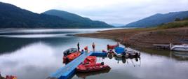 Na zdjęciu widoczny zalew Klimkówka oraz pomost na których stoją strażacy uczestniczący w ćwiczeniach z zakresu ratownictwa wodnego. Do pomostu przycumowane są łódki, ponton i skuter wodny.